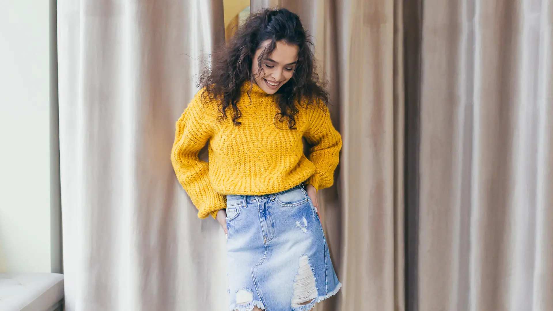 Portrait of a young beautiful woman girl dressed and posing in new clothes in a yellow warm sweater and denim skirt in a clothing store on a background of brown curtains near the fitting room, posing, showing, bragging, looking at the camera, smiling