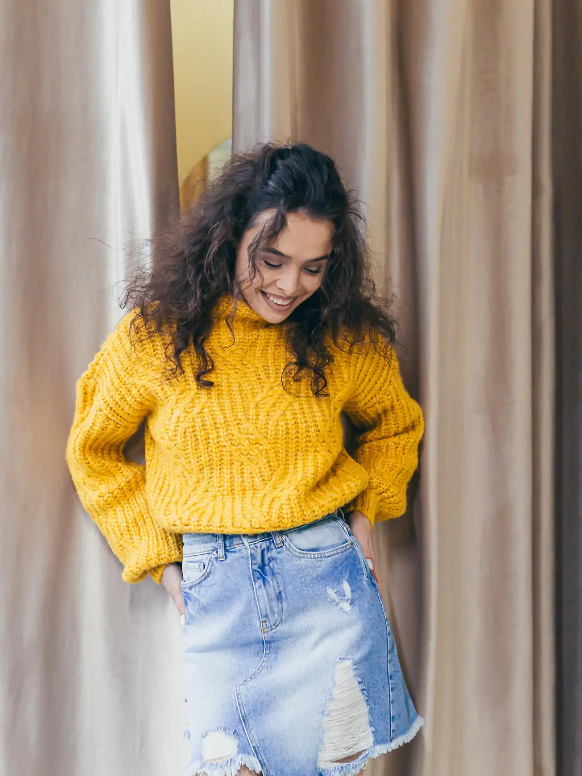 Portrait of a young beautiful woman girl dressed and posing in new clothes in a yellow warm sweater and denim skirt in a clothing store on a background of brown curtains near the fitting room, posing, showing, bragging, looking at the camera, smiling