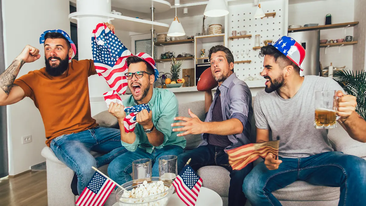 Excited male friends cheering while watching American football match on sofa at home.
