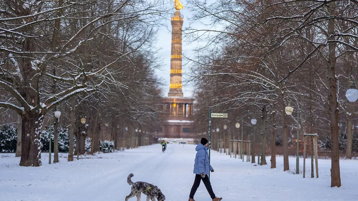 Eine Frau geht mit ihrem Hund vor der Kulisse der Siegessäule im verschneiten Tiergarten spazieren.