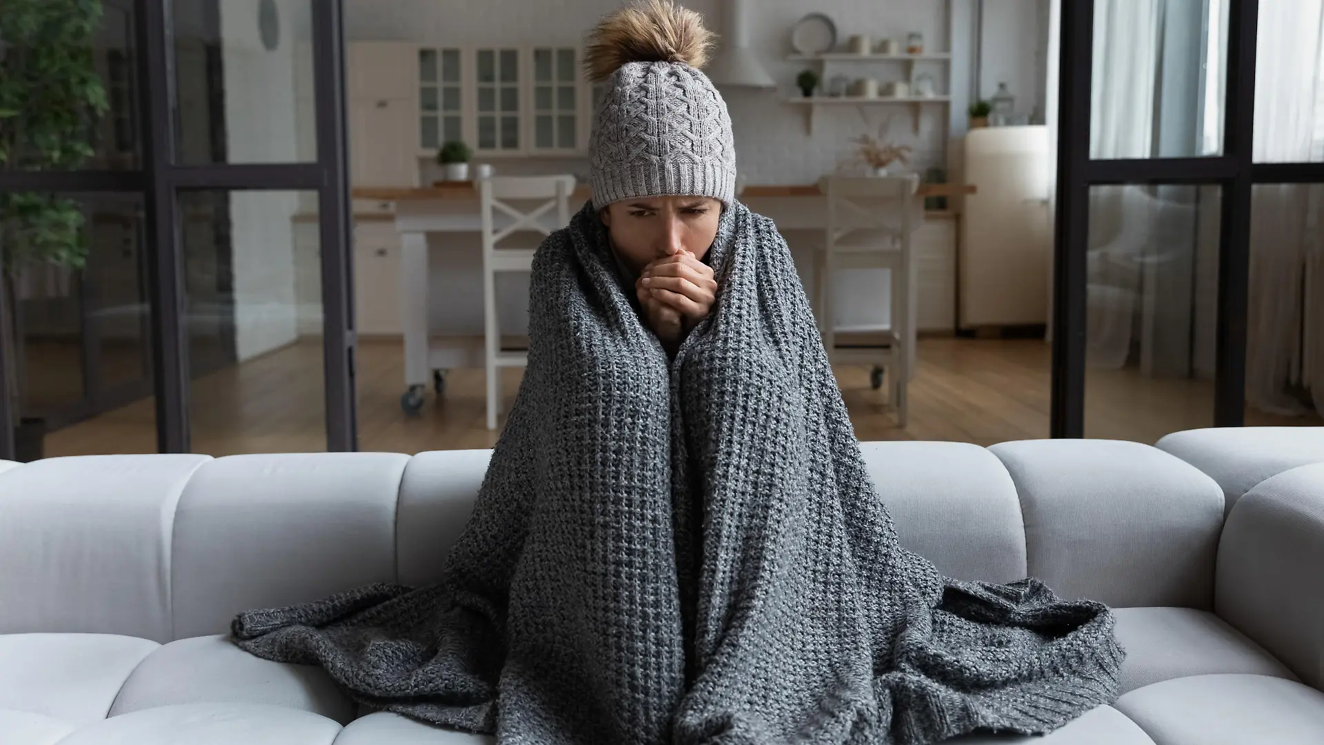 Frozen. Sad latina female sit on couch at freezing cooled studio flat in warm cap and blanket shiver tremble with cold. Unhappy young lady spend time at home feel bad suffer of heating system problems