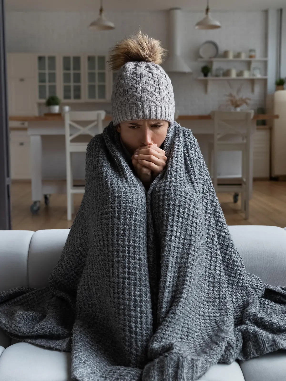 Frozen. Sad latina female sit on couch at freezing cooled studio flat in warm cap and blanket shiver tremble with cold. Unhappy young lady spend time at home feel bad suffer of heating system problems
