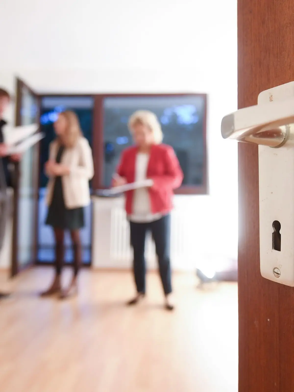 Eine Maklerin spricht mit Interessenten bei einer Wohnungsbesichtigung während im Vordergrund eine Türklinke zu sehen ist.
