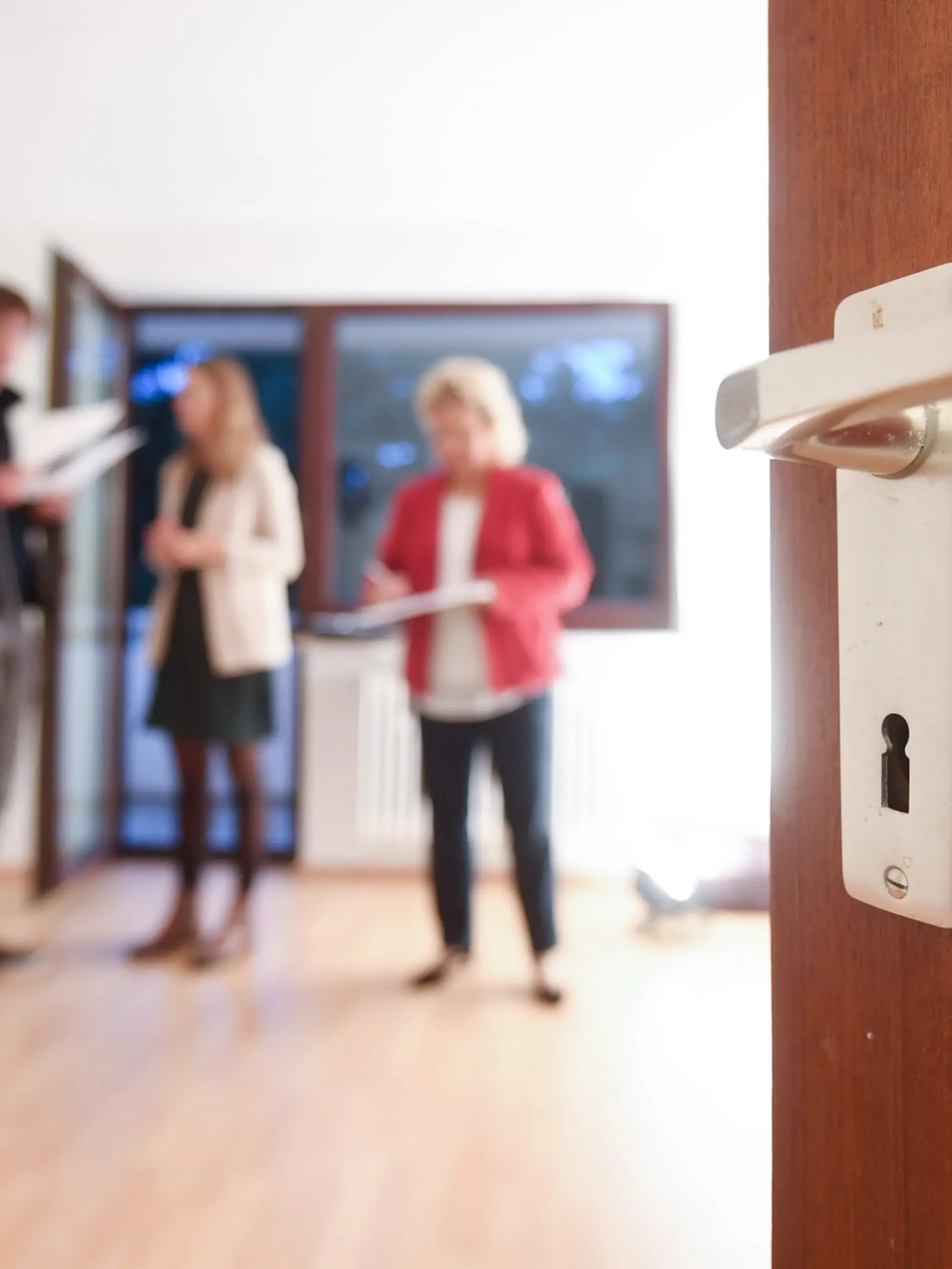 Eine Maklerin spricht mit Interessenten bei einer Wohnungsbesichtigung während im Vordergrund eine Türklinke zu sehen ist.