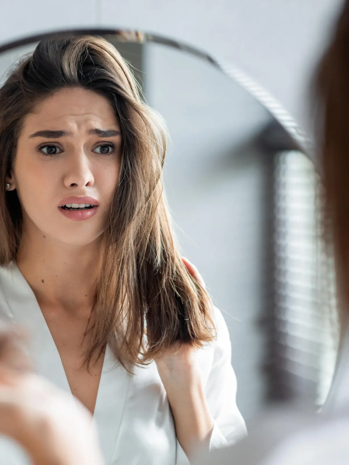Portrait of stressed young woman with bunch of fallen hair in hand looking at mirror in bathroom, scared upset millennial lady in white silk robe suffering hairloss problem, selective focus