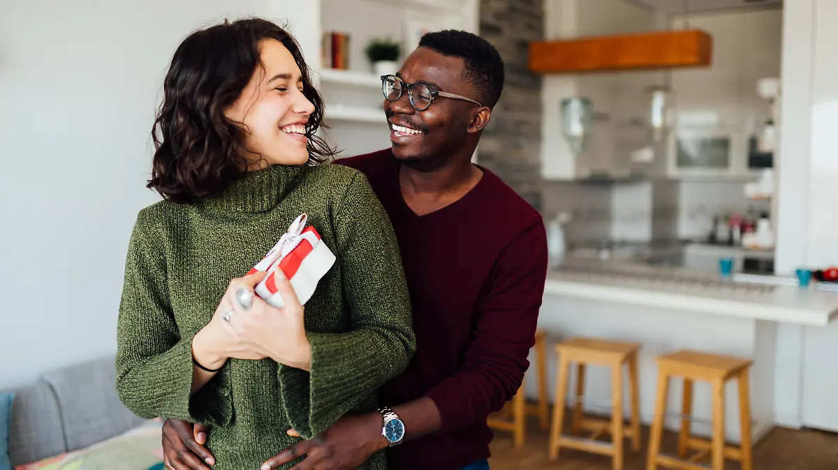 Happy young interracial couple having fun at home, African American boyfriend surprising his Caucasian girlfriend with a Valentine''s day present, making her smile