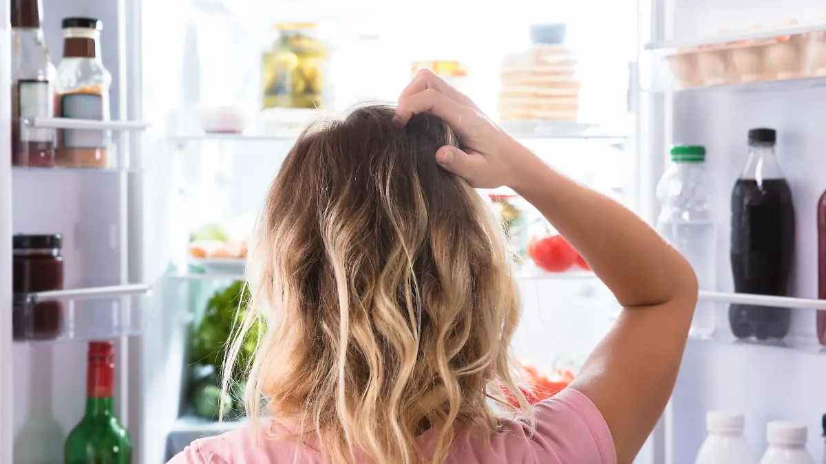 Rear View Of A Confused Woman Looking In Open Refrigerator At Home