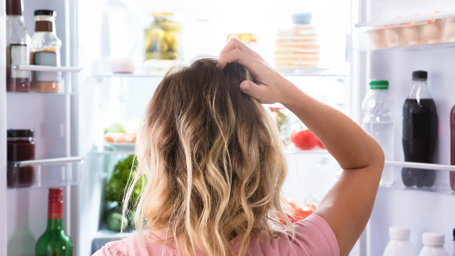 Rear View Of A Confused Woman Looking In Open Refrigerator At Home