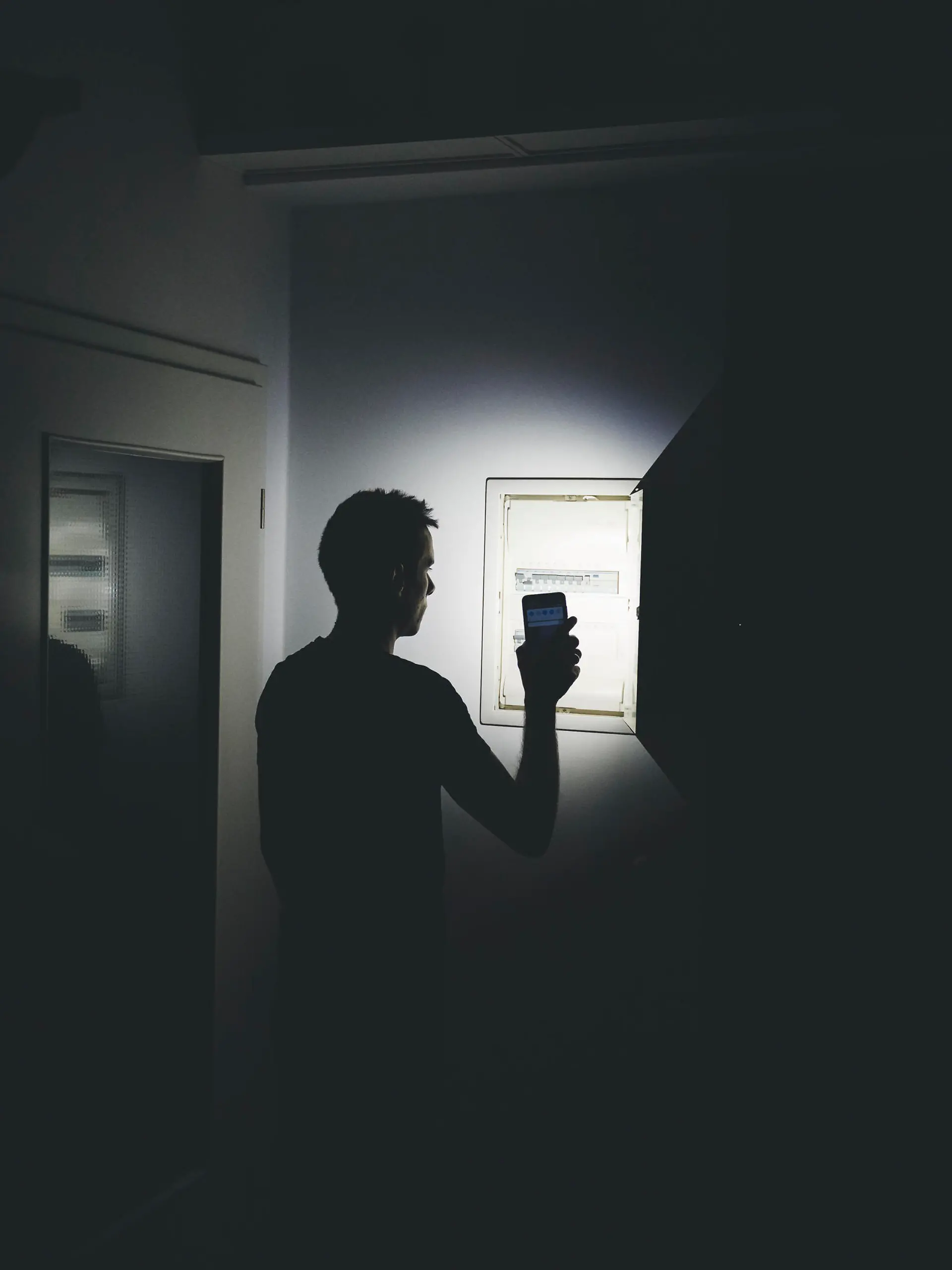 Stromausfall in der Wohnung, Mann leuchtet mit der Taschenlampe in seinem Smartphone in der dunklen Wohnung und überprüft den Sicherungskasten. Elektrizität, Strom Ausfall, Blackout Konzept