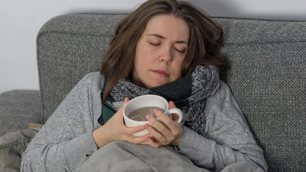 Eine kranke Frau schläft auf der Couch, zugedeckt mit einer Decke und einer Tasse Tee in der Hand || Modellfreigabe vorhanden