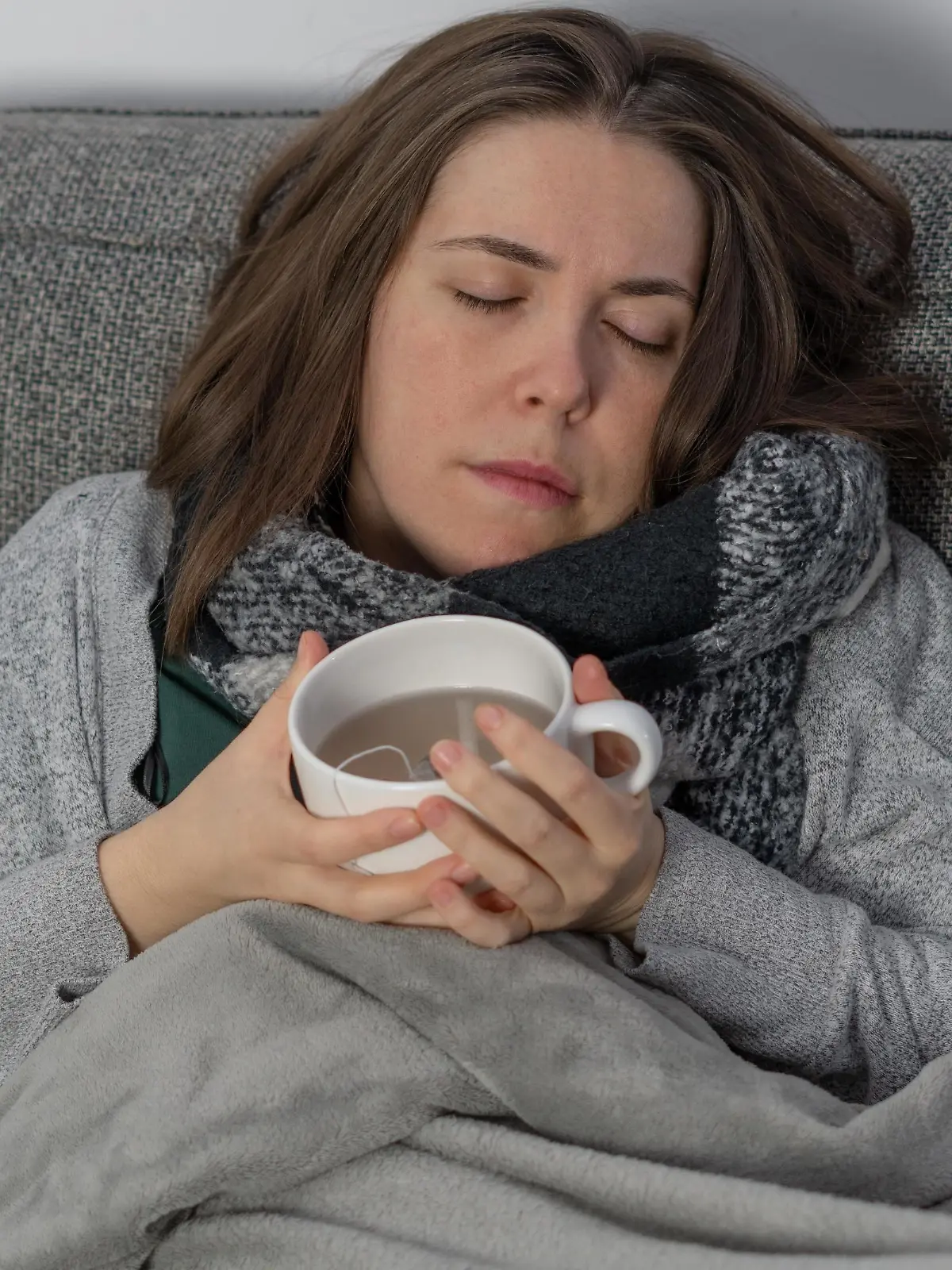 Eine kranke Frau schläft auf der Couch, zugedeckt mit einer Decke und einer Tasse Tee in der Hand || Modellfreigabe vorhanden