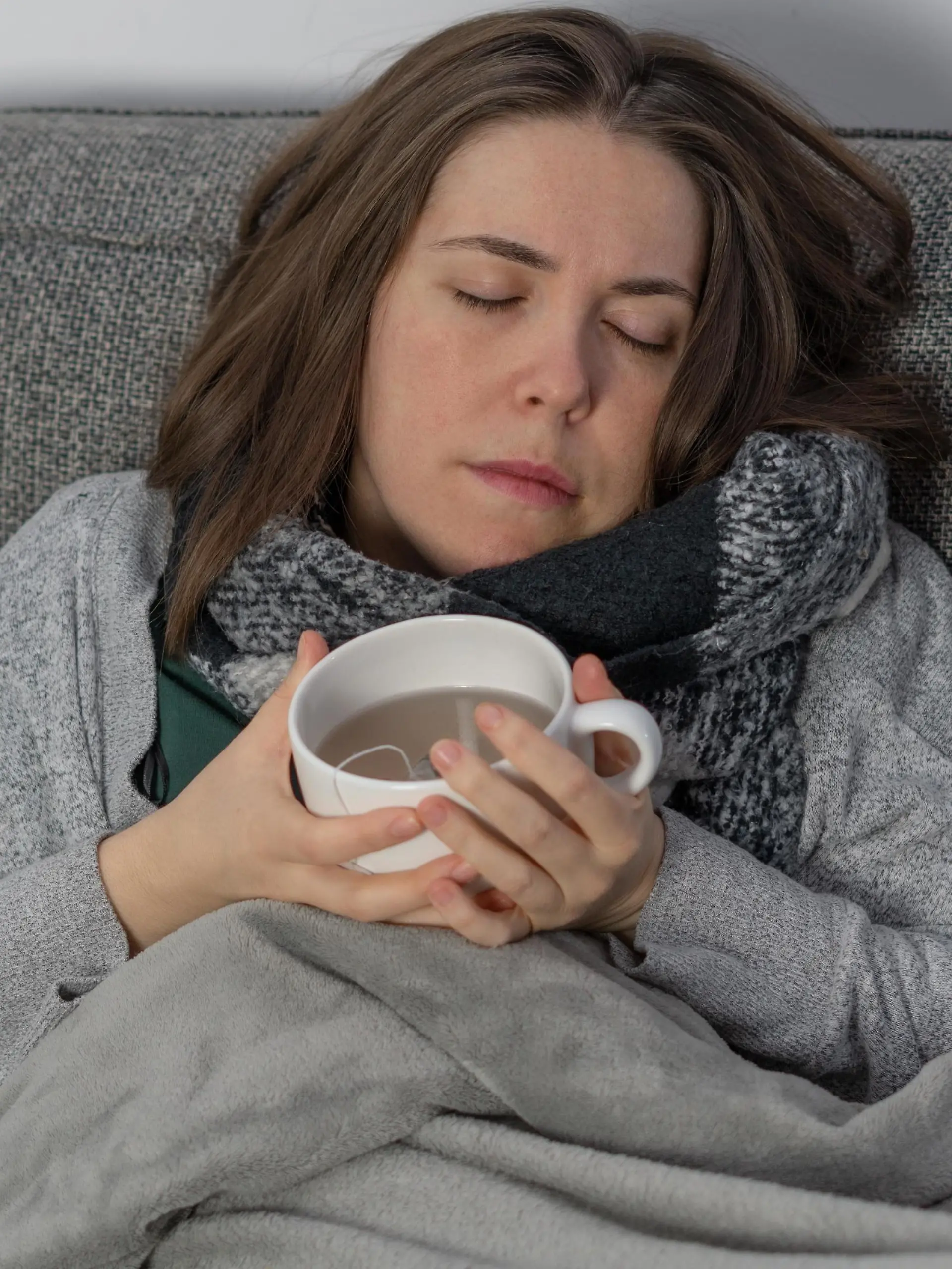 Eine kranke Frau schläft auf der Couch, zugedeckt mit einer Decke und einer Tasse Tee in der Hand || Modellfreigabe vorhanden