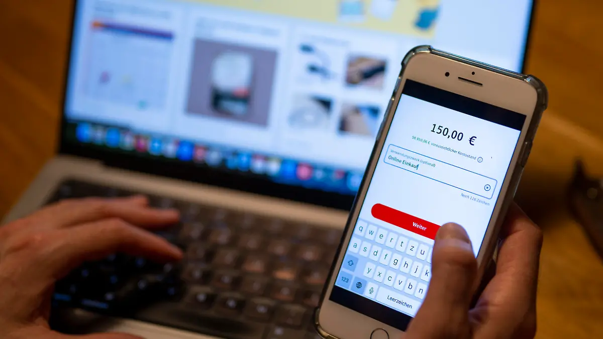 Ein Mann überweist in einer Banking-App auf einem Smartphone Geld, während im Hintergrund auf einem Laptop die Seite eines Online-Handels angezeigt wird.