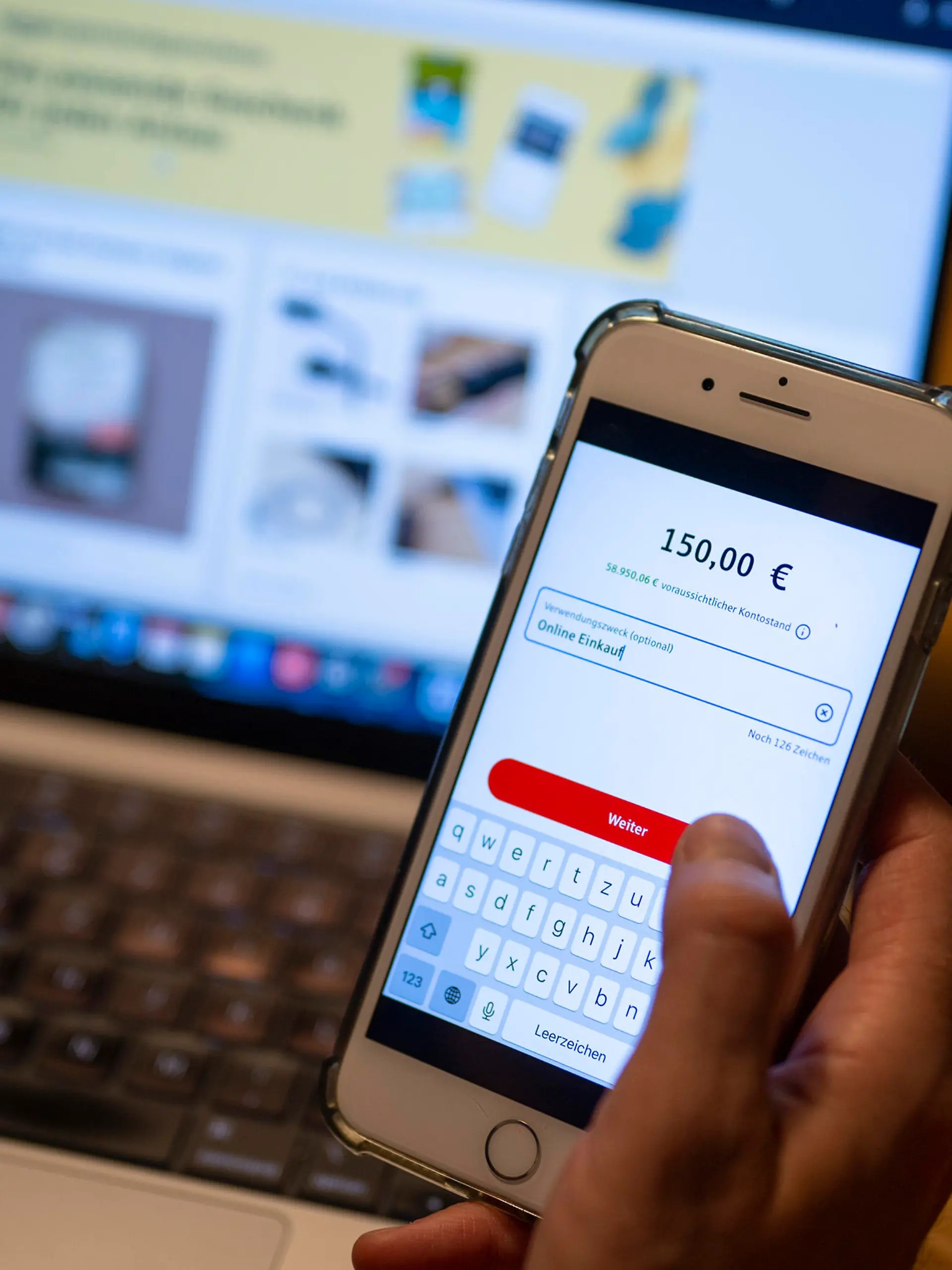 Ein Mann überweist in einer Banking-App auf einem Smartphone Geld, während im Hintergrund auf einem Laptop die Seite eines Online-Handels angezeigt wird.