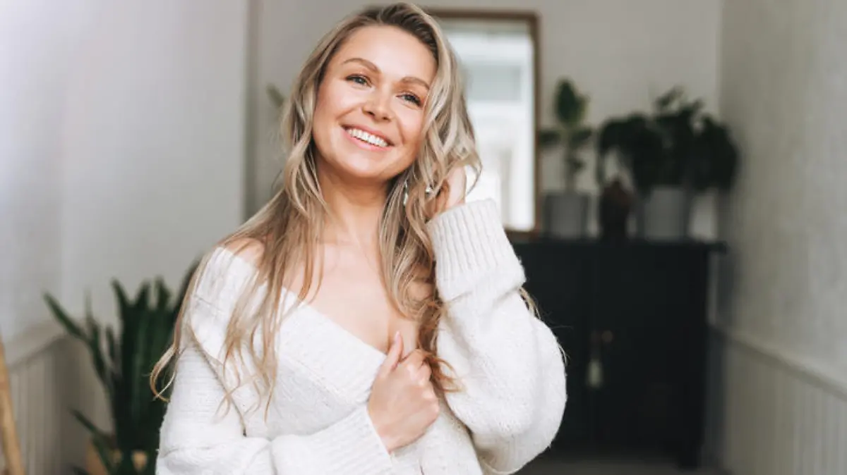 Blonde smiling woman 35 year clean fresh face and hands with long hair in cozy knitted cardigan at the bright interior