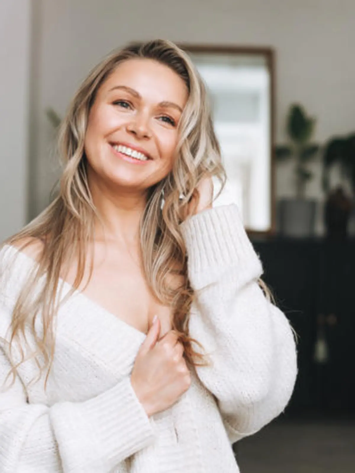 Blonde smiling woman 35 year clean fresh face and hands with long hair in cozy knitted cardigan at the bright interior