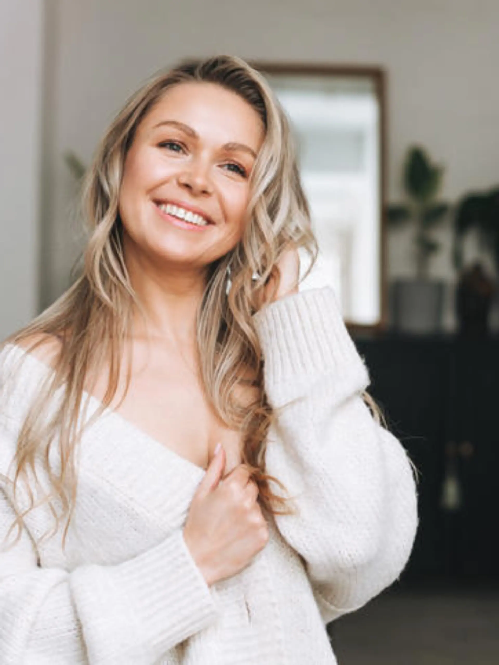 Blonde smiling woman 35 year clean fresh face and hands with long hair in cozy knitted cardigan at the bright interior