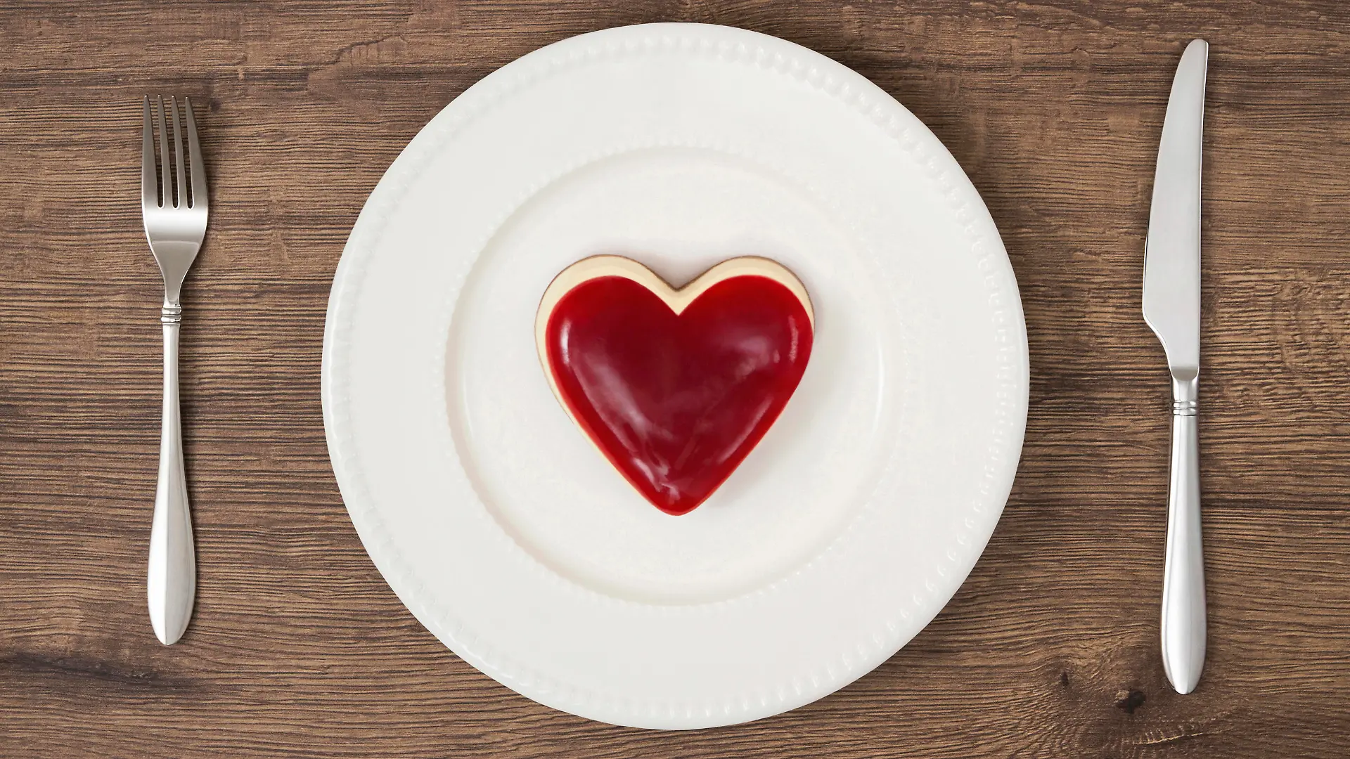 Heart shaped whole mini raspberry cheesecake on red stripe plate with fork and table knife on wooden rustic background.