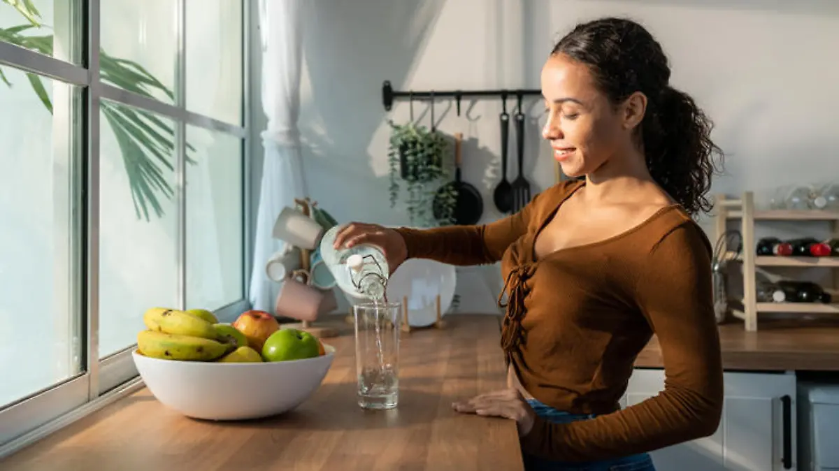 Young beautiful Latino woman pouring clean water into glass in kitchen. Attractive active thirsty girl drink or take a sips of mineral natural in cup for health care and wellbeing in kitchen in house.