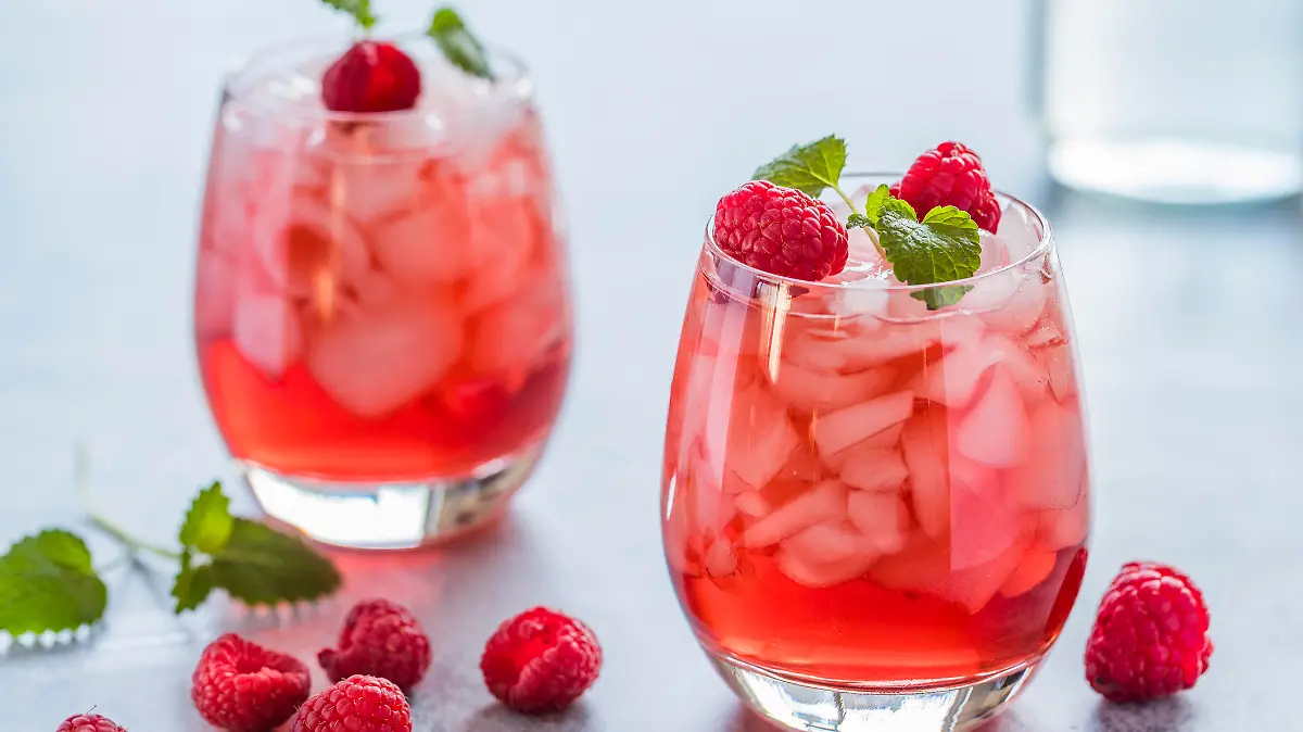 Two refreshing red translucent drinks with ice and raspberries. The raspberry juice is garnished with a green leaf, and there are pieces of ice and raspberries scattered around the drinks.