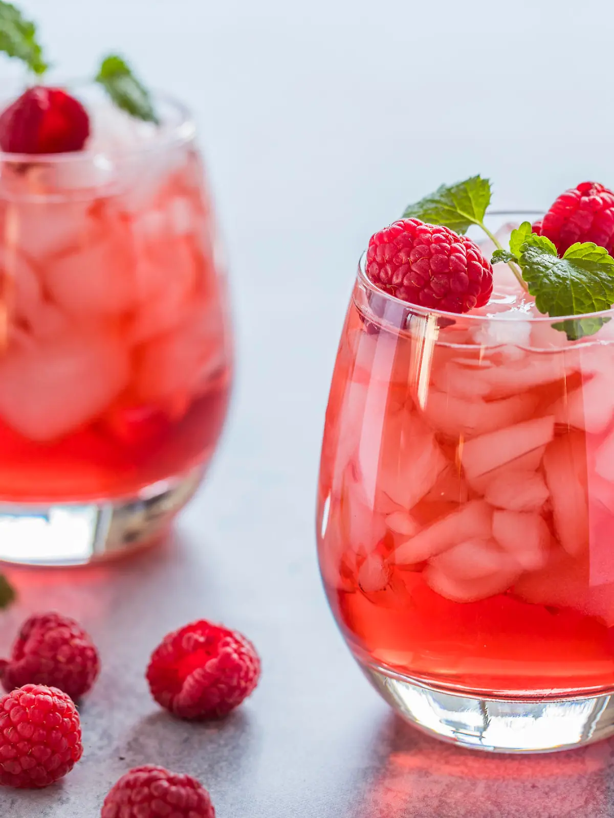 Two refreshing red translucent drinks with ice and raspberries. The raspberry juice is garnished with a green leaf, and there are pieces of ice and raspberries scattered around the drinks.