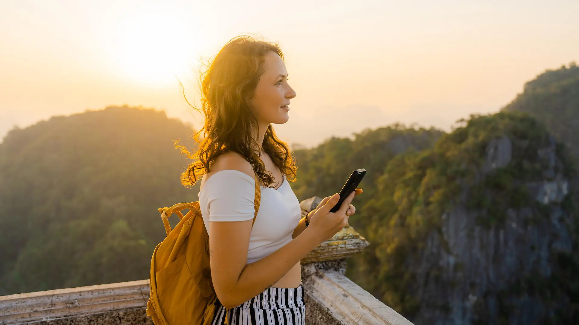 Young Caucasian woman standing on the top of wat Tham Sua in Krabi at sunset and using smartphone