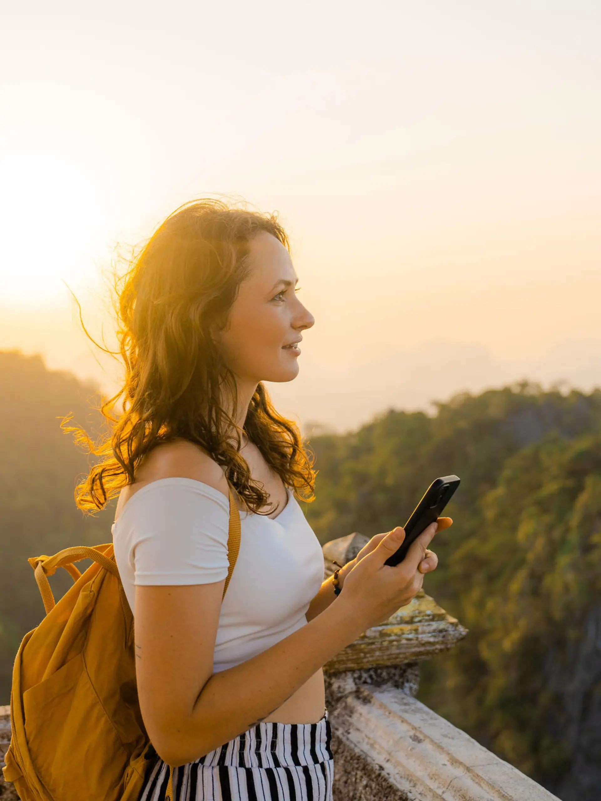 Young Caucasian woman standing on the top of wat Tham Sua in Krabi at sunset and using smartphone