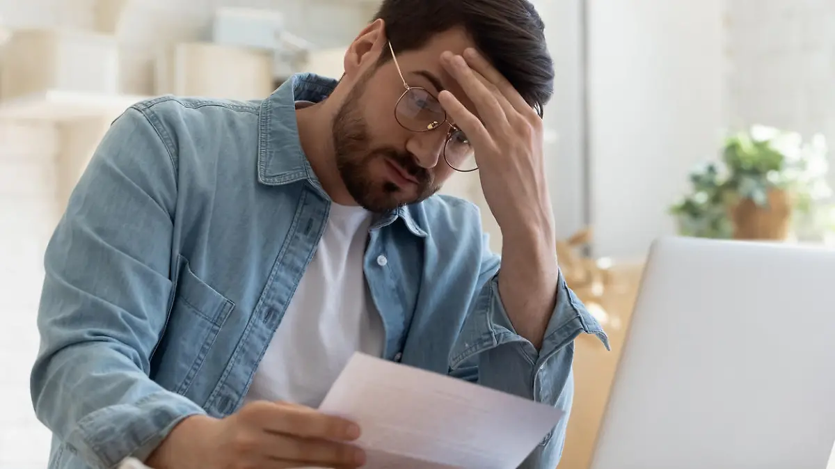 Upset frustrated young man reading bad news in postal mail letter paper document sit at home table, depressed stressed guy worried about high bill tax invoice, overdue debt notification money problem