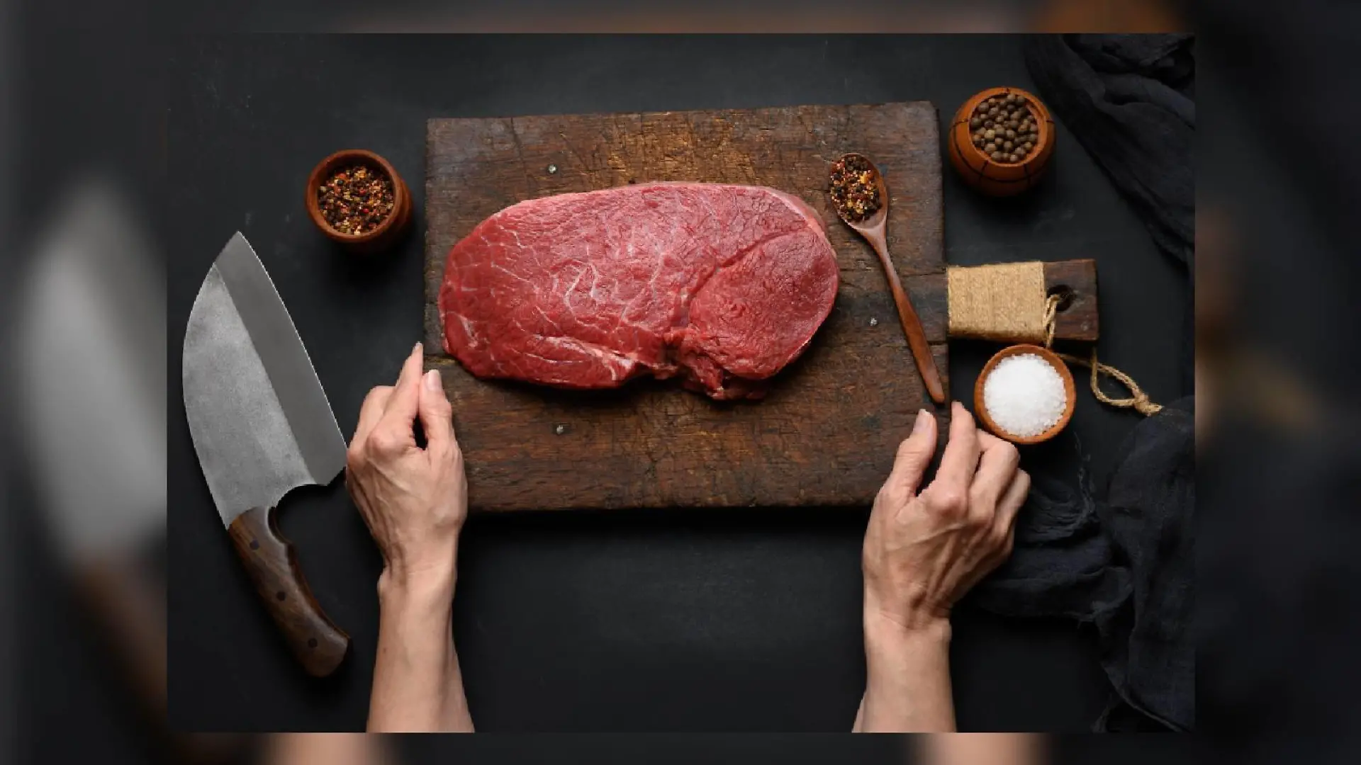 Raw beef tenderloin lies on a wooden cutting board and spices for cooking on a black table, top view and large kitchen knife