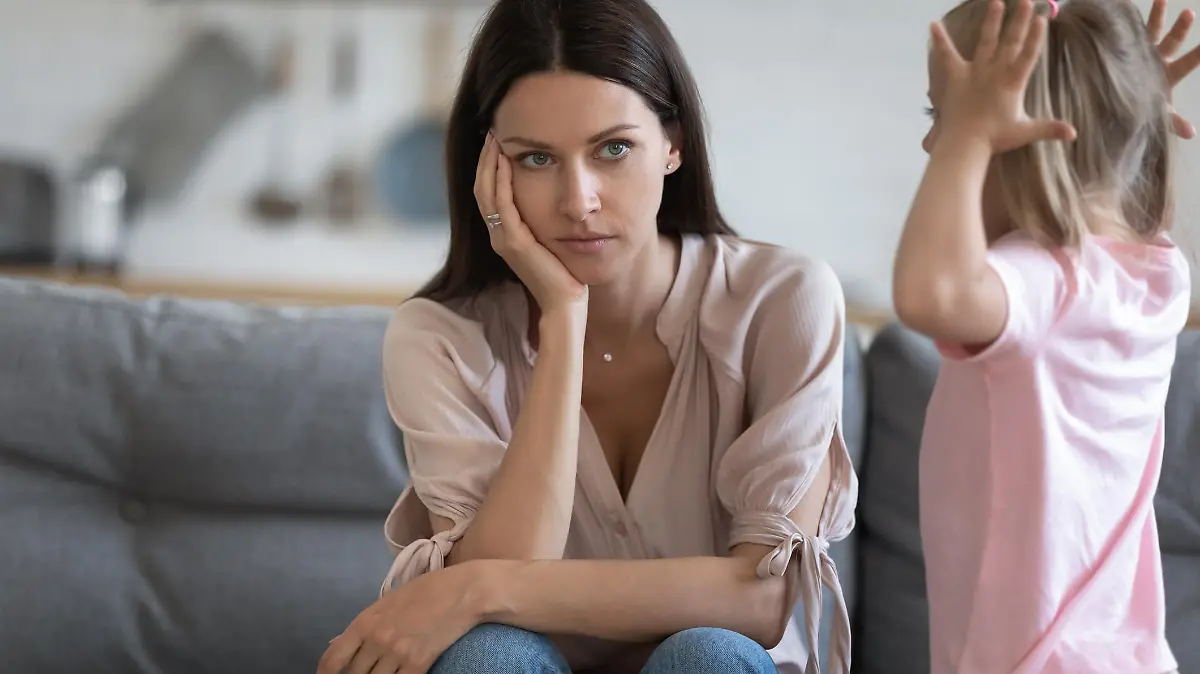 Unhappy mother having problem with noisy naughty little daughter screaming, demanding attention, child tantrum manipulation concept, tired thoughtful mum holding head, sitting on couch at home