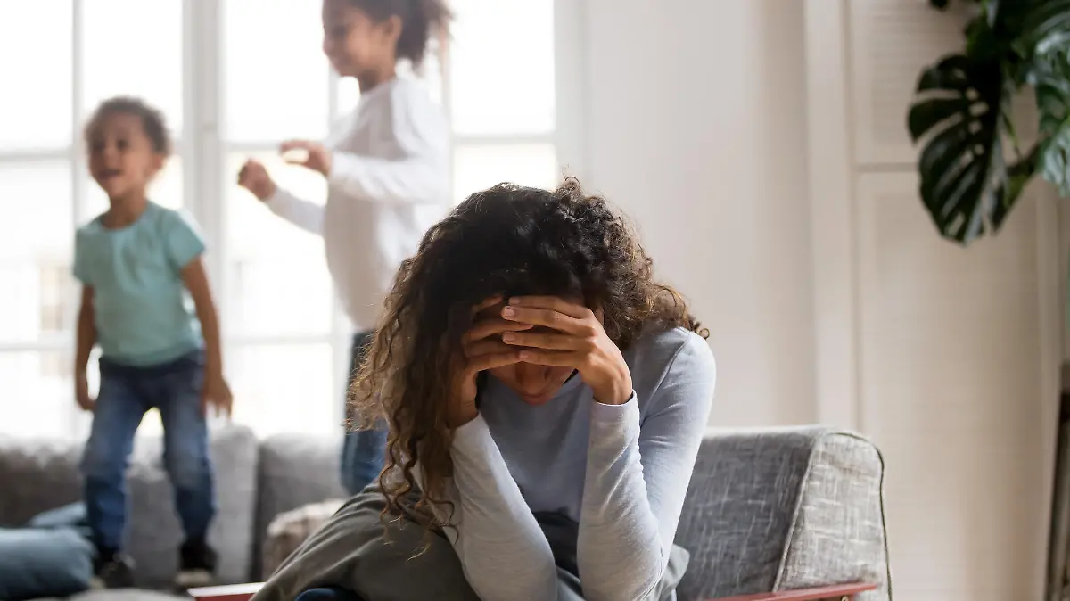 Black single frustrated woman hold her head with hands sitting on chair in living room, playful kids jumping on couch on a background. Tiredness, depression difficult to educate children alone concept