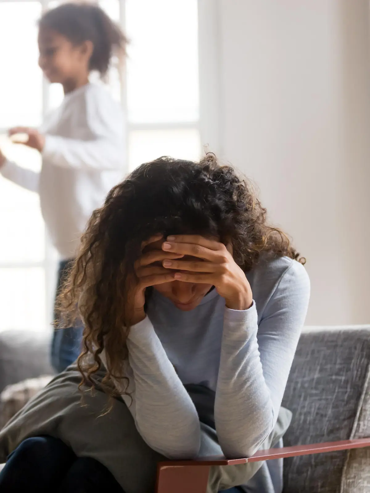 Black single frustrated woman hold her head with hands sitting on chair in living room, playful kids jumping on couch on a background. Tiredness, depression difficult to educate children alone concept