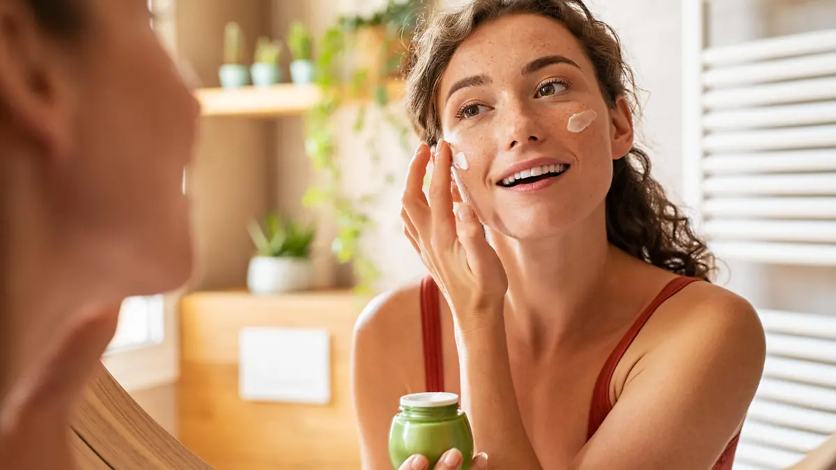 Woman caring of her beautiful skin face standing near mirror in the bathroom. Young woman applying moisturizing cream on her face during morning routine. Smiling natural girl holding little green jar of ecological cosmetic cream.