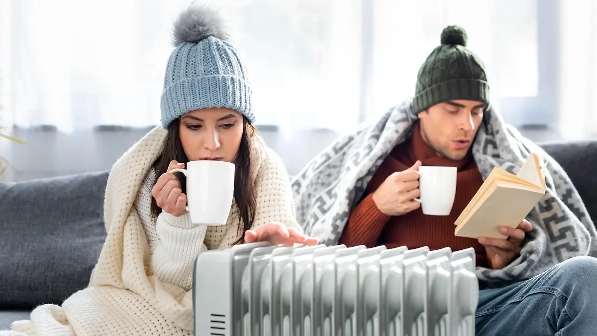 attractive girlfriend with cup of tea and boyfriend reading book and warming up near heater