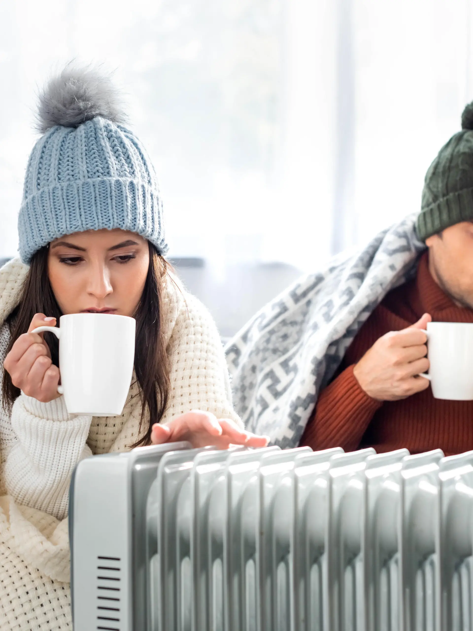 attractive girlfriend with cup of tea and boyfriend reading book and warming up near heater