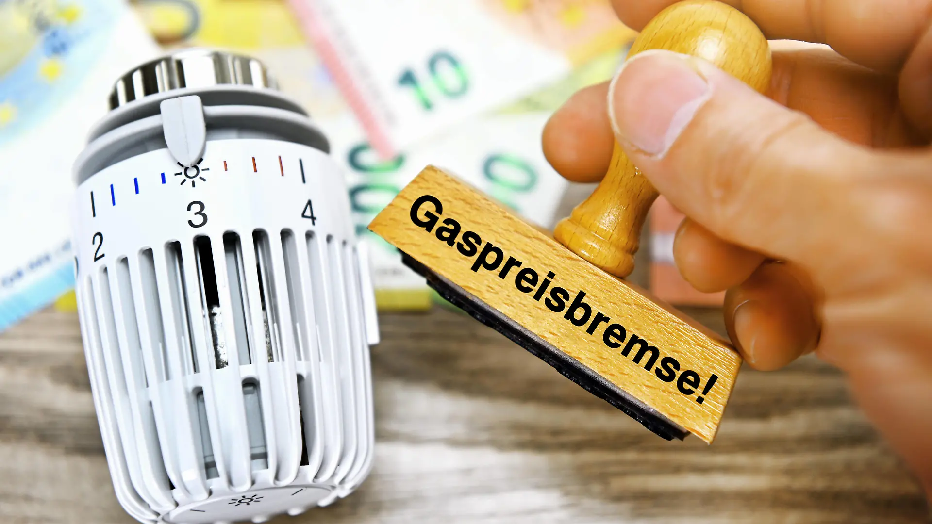FOTOMONTAGE, Hand hält Stempel mit Aufschrift Gaspreisbremse neben Heizungsthermostat *** PHOTOMONTAGE, hand holding stamp with inscription gas price brake next to heating thermostat