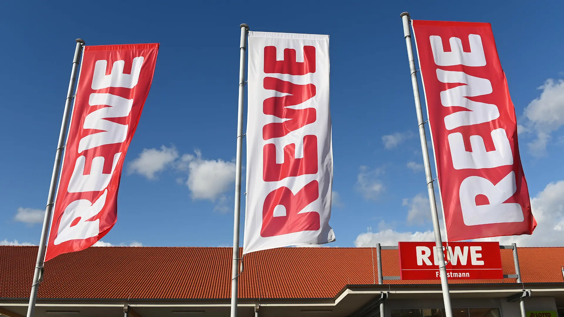 ARCHIV - 25.02.2020, Hessen, Volkmarsen: Fahnen mit dem Logo des Lebensmitteleinzelhändlers Rewe wehen vor einer Filiale. Ein Ende der Preiserhöhungen im deutschen Lebensmittelhandel ist nach Einschätzung von Rewe-Chef Souque nicht in Sicht. (zu dpa "Rewe-Chef rechnet mit weiteren Preissteigerungen") Foto: Uwe Zucchi/dpa +++ dpa-Bildfunk +++