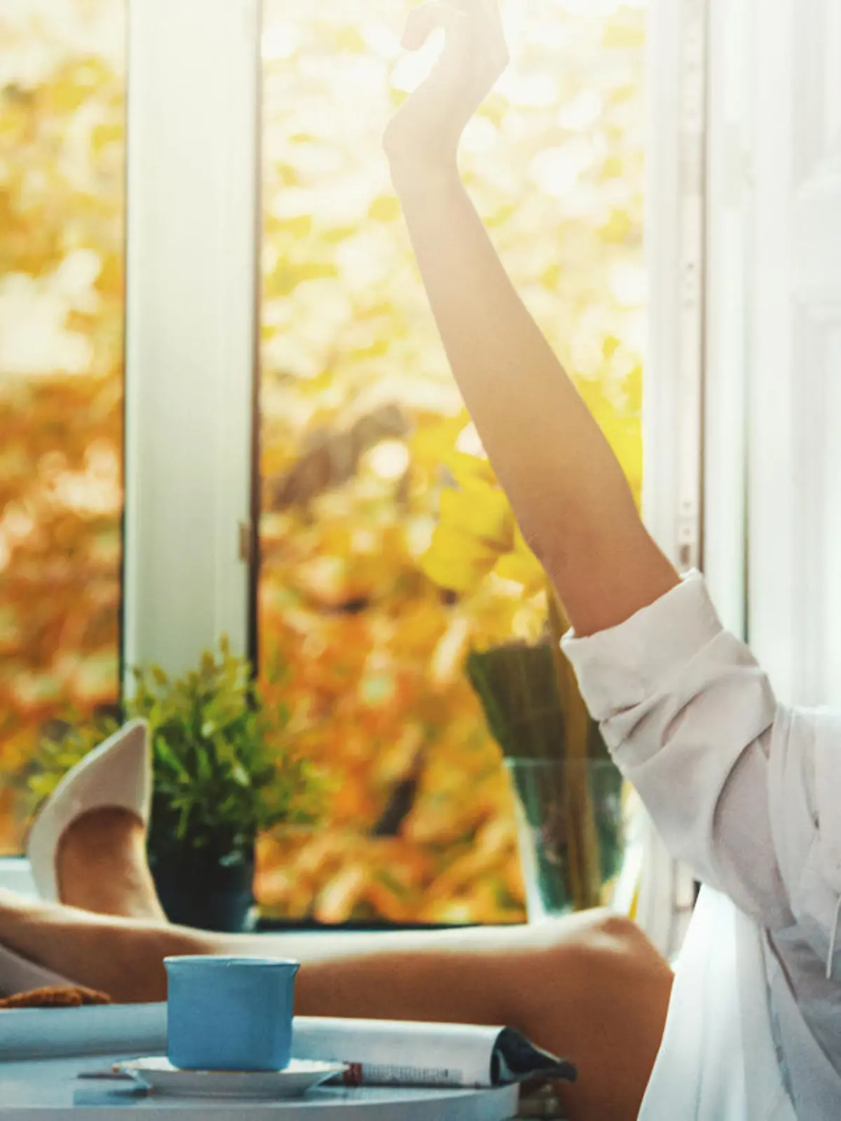 Closeup rear view of attractive yet unrecognizable blond woman sitting next to her bedroom window wearing only white men's shirt and high heels. It's beautiful day outside and she's stretching her arms but she is so lazy at the moment. There is a cup of coffee and a magazine on the night stand next to her.