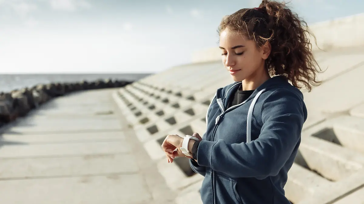 Young purposeful curly-haired fitness woman in sportswear looking at watch on city beach