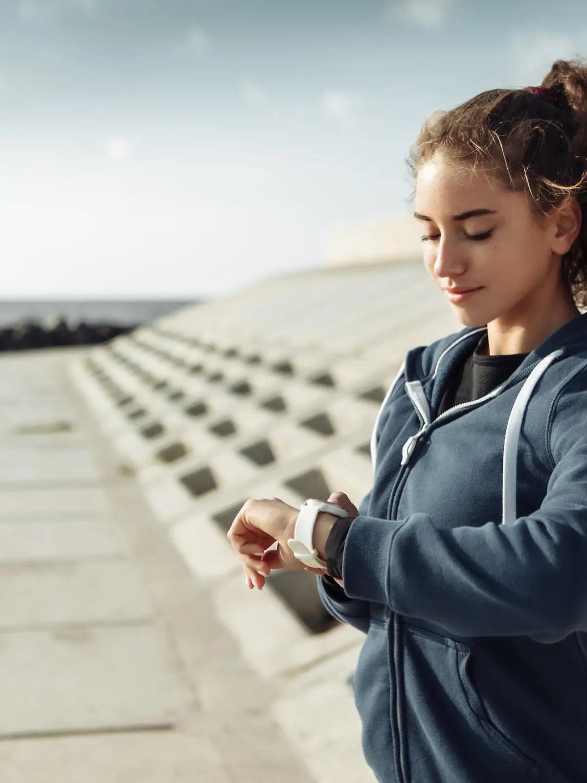 Young purposeful curly-haired fitness woman in sportswear looking at watch on city beach